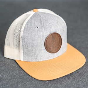 May include: A grey and yellow baseball cap with a white mesh back. The cap has a brown leather patch with an eagle design and the text "1776 2023". The brim is yellow, and the top button is brown.