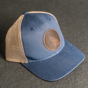 May include: A blue and brown baseball cap with a brown leather patch featuring a wolf silhouette. The cap has a mesh back.