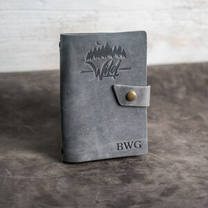 May include: Gray leather journal with a flap closure and a brass button. The front cover features an embossed design of trees and the word "Wild". The journal is personalized with the initials "BWG".
