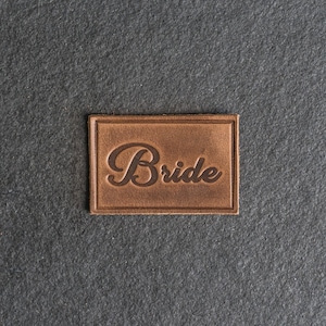 May include: A brown leather patch with the word "Bride" embossed in a cursive font.