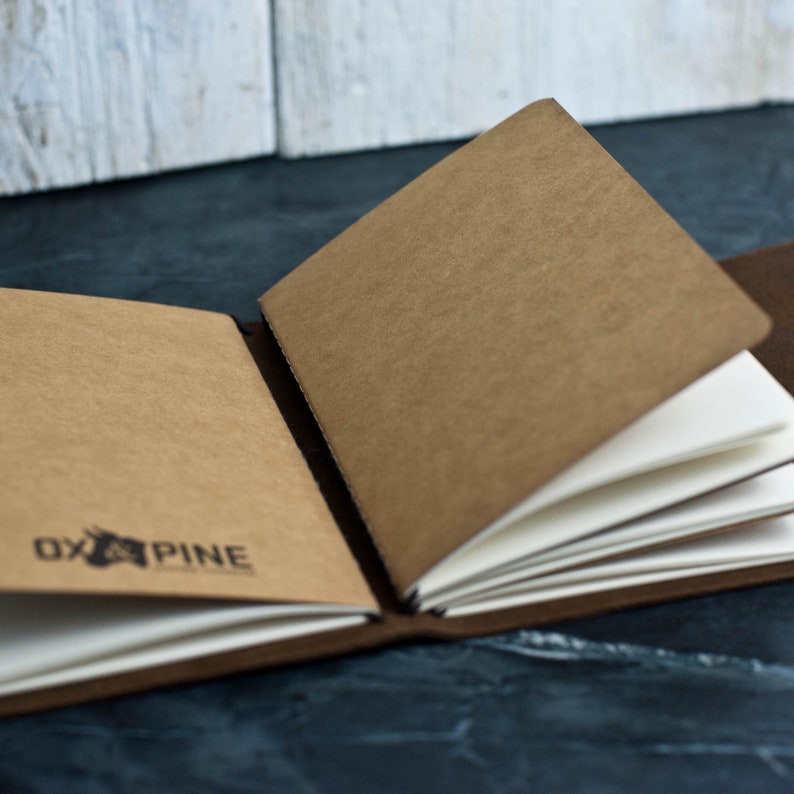 May include: A brown leather-bound journal with blank pages, open to reveal the white paper inside. The cover has the text "Ox & Pine" printed on it.