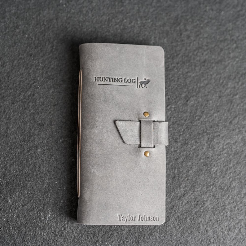 May include: Gray leather hunting log with the text "HUNTING LOG" and a deer silhouette. The journal has a strap closure with brass rivets and the name "Taylor Johnson" embossed at the bottom.