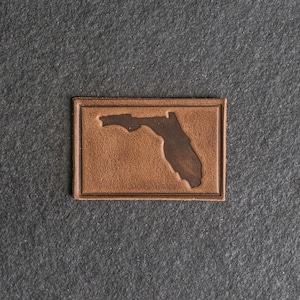 Puede incluir: Un parche de cuero marrón con un contorno marrón oscuro del estado de Florida.