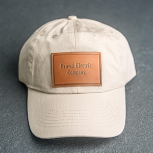 Puede incluir: Una gorra de béisbol de color beige con un parche rectangular de cuero marrón claro centrado en la parte delantera. El parche tiene grabado el texto Brown Electric Company en una fuente serif. La gorra tiene visera curva y paneles cosidos.