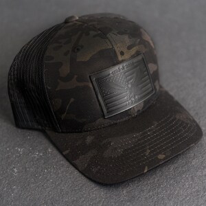 May include: A black camouflage baseball cap with a mesh back. The front features a black leather patch with an American flag design and the text "1776-2024". The cap has a curved brim and a button on top.