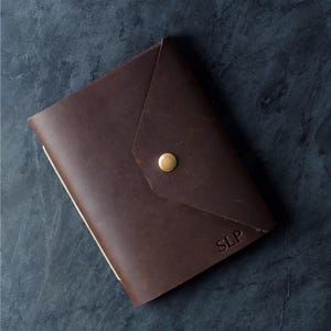 May include: A dark brown leather journal cover featuring a triangular flap closure secured with a single brass-colored metal snap button. The bottom right corner of the cover is embossed with the capital letters SLP in a simple serif font.