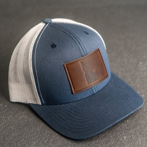 Puede incluir: Una gorra de camionero azul y blanca con un parche de cuero marrón que presenta el contorno del estado de Idaho.