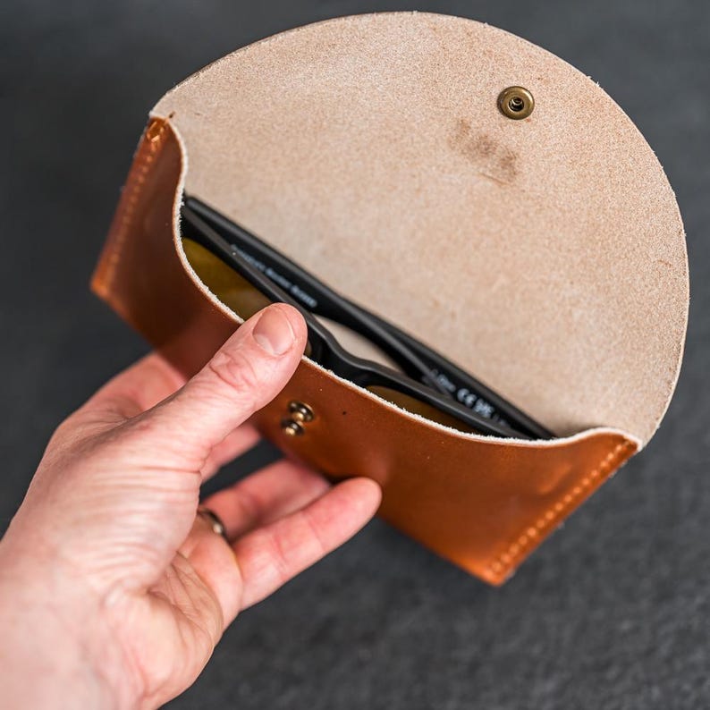 Puede incluir: Un estuche de cuero marr&oacute;n para gafas de sol, abierto para revelar un par de gafas de sol. El estuche se mantiene abierto con una mano, mostrando el forro interior y un cierre de bot&oacute;n a presi&oacute;n de lat&oacute;n. El estuche est&aacute; hecho de cuero cosido.