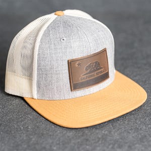 May include: A grey and yellow trucker hat with a white mesh back. The front panel has a brown leather patch with the California Republic flag and the text "CALIFORNIA REPUBLIC". The hat has a curved peak and a button on top.