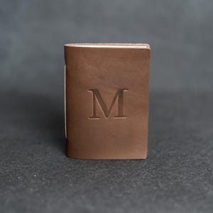 May include: A brown leather wallet with the letter "M" embossed on the front. The wallet is rectangular and appears to be closed. The background is a dark gray.