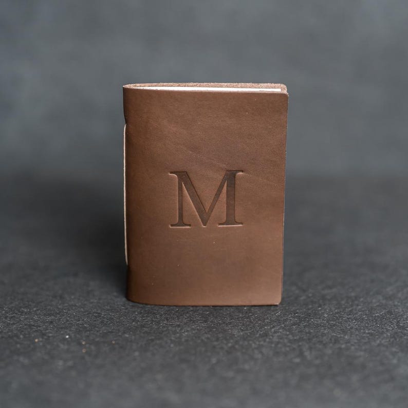 May include: A brown leather wallet with the letter "M" embossed on the front. The wallet is rectangular and appears to be closed. The background is a dark gray.