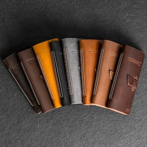 May include: A collection of leather journals in various colors, including brown, black, gray, and orange. Each journal has a stitched spine and the word "Hunting" embossed on the cover. Some journals have additional decorative elements.