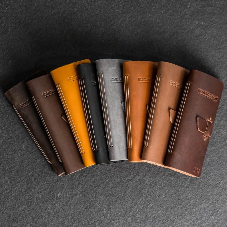 May include: A collection of leather journals in various colors, including brown, black, gray, and orange. Each journal has a stitched spine and the word "Hunting" embossed on the cover. Some journals have additional decorative elements.