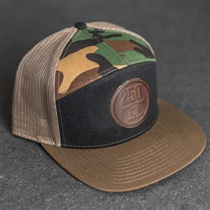 Op de afbeelding: Een pet met een bruine klep en een zwart voorpaneel met een leren patch met de tekst "250 Anniversary". De bovenkant van de pet heeft een camouflagepatroon en de achterkant is een beige mesh.