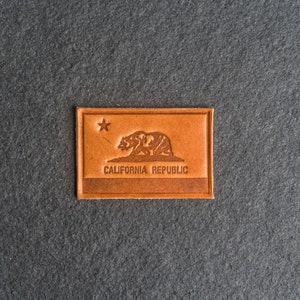 May include: A brown leather patch with the California state flag design. The flag features a grizzly bear, a star, and the text "CALIFORNIA REPUBLIC".