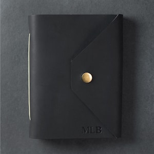 May include: A black leather journal with a triangular flap closure secured by a single brass-colored metal stud. The front cover features the embossed letters MLB in the bottom right corner, and the spine shows light-colored paper edges.