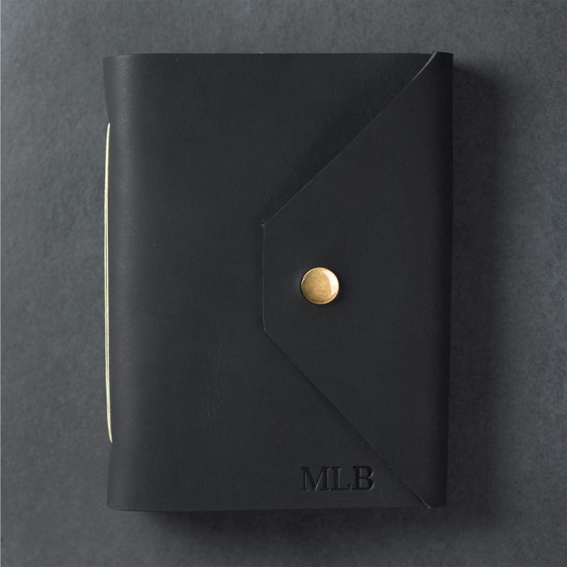 May include: A black leather journal with a triangular flap closure secured by a single brass-colored metal stud. The front cover features the embossed letters MLB in the bottom right corner, and the spine shows light-colored paper edges.