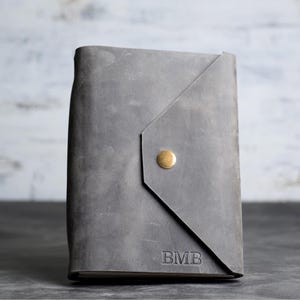 May include: A grey leather journal cover featuring a triangular flap closure secured with a single brass-colored metal snap button. The front surface displays the embossed letters BMB in the bottom right corner, showcasing a matte finish.