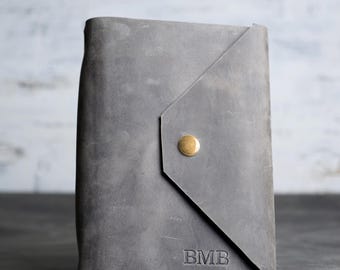 Op de afbeelding: Een grijze lederen omslag voor een notitieboek met een driehoekige flapsluiting, beveiligd met een enkele messingkleurige metalen drukknoop. Het vooroppervlak toont de in reliëf aangebrachte letters BMB in de rechterbenedenhoek.