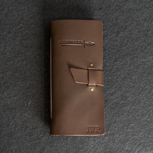 May include: A brown leather fishing log with the text "FISHING LOG" and a fish graphic embossed on the cover. The log has a strap closure with brass rivets and the initials "JWP" embossed at the bottom.