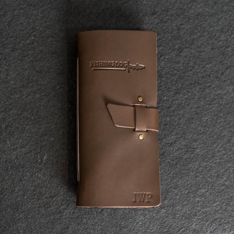 May include: A brown leather fishing log with the text "FISHING LOG" and a fish graphic embossed on the cover. The log has a strap closure with brass rivets and the initials "JWP" embossed at the bottom.