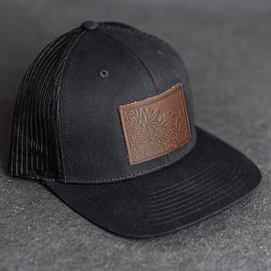 Op de afbeelding: Zwarte truckerpet met een gebogen klep en een mesh achterkant. Op het voorpaneel zit een bruine leren patch met een reliëf bloemenontwerp. De pet heeft een verstelbare snapback sluiting.
