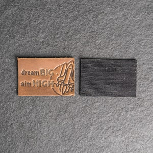 May include: Brown leather patch with the text "dream BIG aim HIGH" and a graphic of a rocket ship. The patch is attached to a black hook and loop fastener.