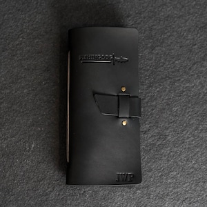 May include: Black leather fishing log with the text "FISHING LOG" embossed on the front. The log has a strap closure with brass rivets and the initials "JWP" embossed at the bottom. The log is closed.