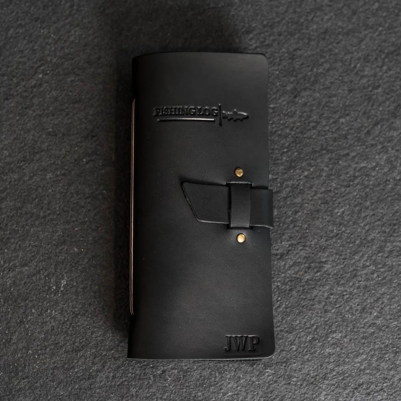 May include: Black leather fishing log with the text "FISHING LOG" embossed on the front. The log has a strap closure with brass rivets and the initials "JWP" embossed at the bottom. The log is closed.