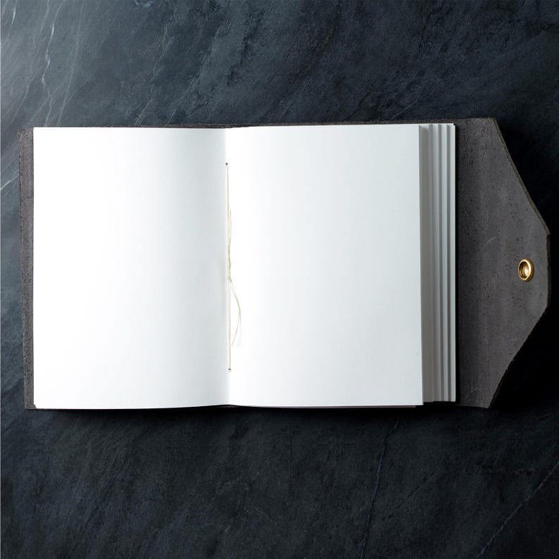 May include: An open journal with a dark charcoal gray leather cover and a pointed flap closure secured by a single brass stud. The interior features blank white pages bound with visible light-colored thread along the center spine fold.