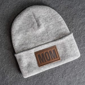 May include: A gray knit beanie with a brown leather patch that reads "MOM". The hat is folded at the brim and is made of a soft, textured material. The patch is rectangular and has the word "MOM" embossed on it.