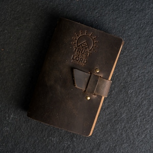 May include: A brown leather journal with a strap closure. The cover features an embossed design of a mountain with a sun and the text "TRUST IN THE LORD, Proverbs 3:5-6".