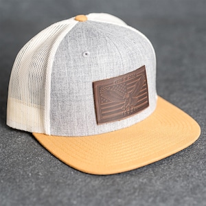 May include: Grey and white trucker cap with a yellow peak. The cap features a brown leather patch with an American flag design and the text "1776 2016". It has a mesh back and a button on top.