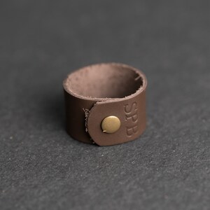 Op de afbeelding: Een bruine leren manchetarmband met een ronde koperen drukknoopsluiting en de in reli&euml;f aangebrachte letters SPB op de buitenkant. De band is gemaakt van &eacute;&eacute;n stuk leer met een zichtbare nerf en een glad, afgerond ontwerp aan de randen.