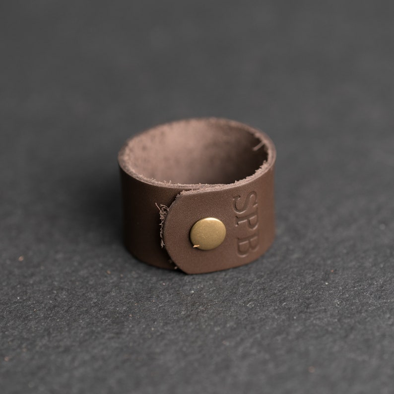 Op de afbeelding: Een bruine leren manchetarmband met een ronde koperen drukknoopsluiting en de in reli&euml;f aangebrachte letters SPB op de buitenkant. De band is gemaakt van &eacute;&eacute;n stuk leer met een zichtbare nerf en een glad, afgerond ontwerp aan de randen.