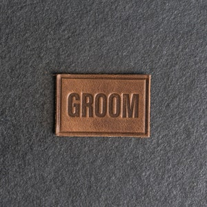 Puede incluir: Un parche de cuero marrón con la palabra "GROOM" en relieve en marrón oscuro.