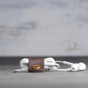 May include: White wired earbuds coiled and secured with a dark brown leather cable organizer. The leather strap features a gold-toned metal snap button and the embossed letters KWP. The earbuds have a standard 3.5mm jack and a small inline remote.