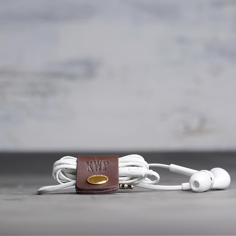 May include: White wired earbuds coiled and secured with a dark brown leather cable organizer. The leather strap features a gold-toned metal snap button and the embossed letters KWP. The earbuds have a standard 3.5mm jack and a small inline remote.