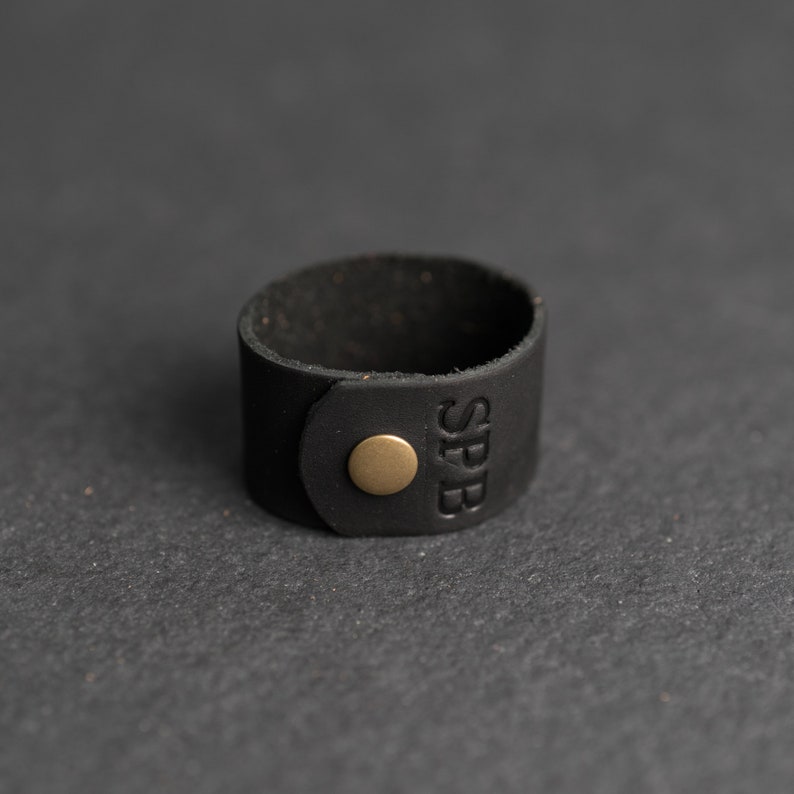 Op de afbeelding: Een zwarte leren manchetarmband met een gladde matte afwerking en een ronde, messingkleurige metalen drukknoopsluiting. De letters SPB zijn verticaal in het leeroppervlak bij de sluiting gedrukt, wat zorgt voor een eenvoudig ontwerp.
