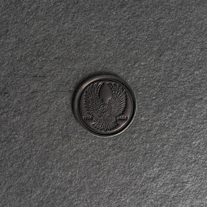 May include: A dark gray, circular emblem with a detailed eagle design, wings spread, centered within a border. The emblem is embossed with the years 1975 and 2025. The background is a textured, dark gray surface.