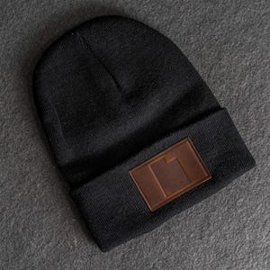 May include: A black knit beanie with a brown rectangular patch on the front. The patch has a geometric design. The beanie is made of a soft, warm material, perfect for cold weather. The hat is a versatile accessory.