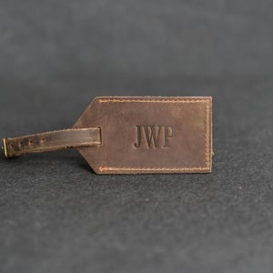May include: A brown leather luggage tag with the initials "JWP" embossed on the front. The tag has a stitched border and a small strap with a buckle. The background is a dark gray.
