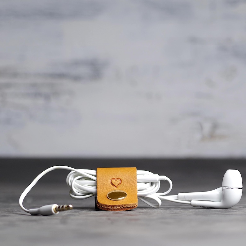 May include: A tan leather cord organizer with a small embossed heart and a gold-toned metal snap button, wrapped around a coiled white wired earphone set with a standard 3.5mm jack and two white earbud pieces.