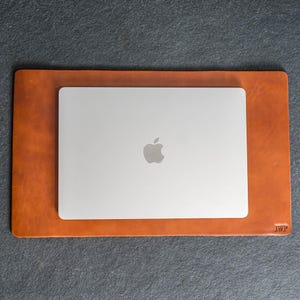 May include: A silver laptop with the Apple logo rests on a rectangular, tan leather desk mat. The mat has rounded corners and a debossed "JWP" monogram in the lower right corner. The mat is approximately 15 inches long and 10 inches wide.