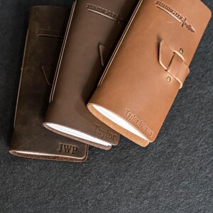 May include: Three leather journals in varying shades of brown. The journals have the words "FISHING LOG" embossed on the top and the name "Taylor Johnson" on one. One journal has the initials "JWP". Each journal has a strap closure.