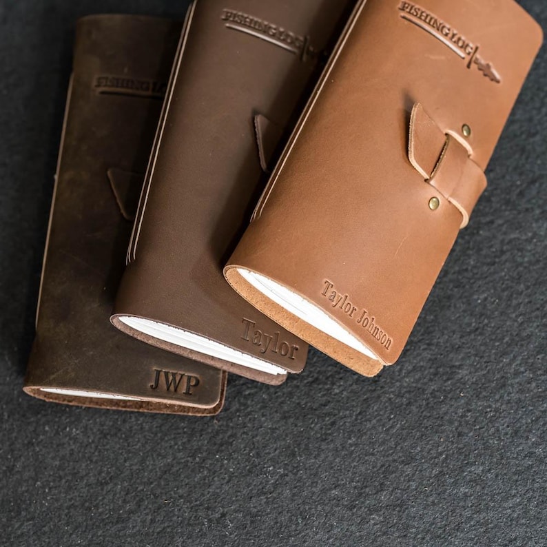 May include: Three leather journals in varying shades of brown. The journals have the words "FISHING LOG" embossed on the top and the name "Taylor Johnson" on one. One journal has the initials "JWP". Each journal has a strap closure.
