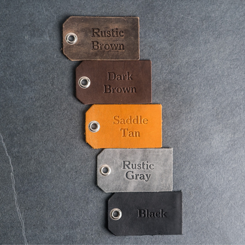 May include: Five rectangular leather tags arranged vertically, each featuring a silver metal eyelet. The tags are labeled with embossed text: Rustic Brown, Dark Brown, Saddle Tan, Rustic Gray, and Black, representing the available color options.