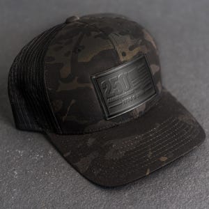 May include: A black camouflage baseball cap with a mesh back. The front of the cap features a black rectangular patch with the text "250 Anniversary 1776-2026" and an American flag design.