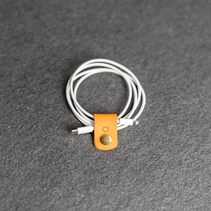 May include: A white charging cable coiled and secured with a tan leather cord organizer. The organizer features a small embossed heart icon and a circular metallic snap button fastener to keep the cable neatly bundled and tangle-free.