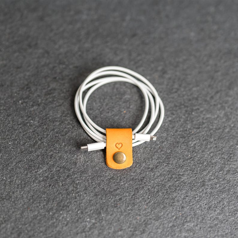 May include: A white charging cable coiled and secured with a tan leather cord organizer. The organizer features a small embossed heart icon and a circular metallic snap button fastener to keep the cable neatly bundled and tangle-free.